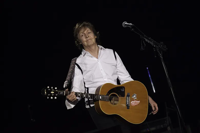 La imagen muestra a Paul McCartney tocando una guitarra acústica en vivo