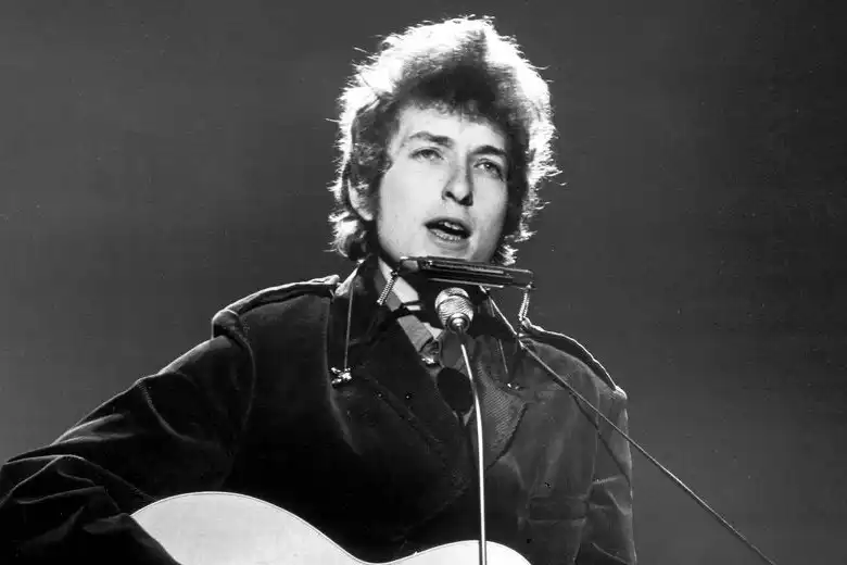 Es una imagen en blanco negro que muestra a Bob Dylan cantando sobre el escenario en los años 60.