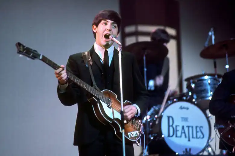 La imagen muestra a Paul McCartney tocando su bajo Höfner y cantando sobre el escenario.